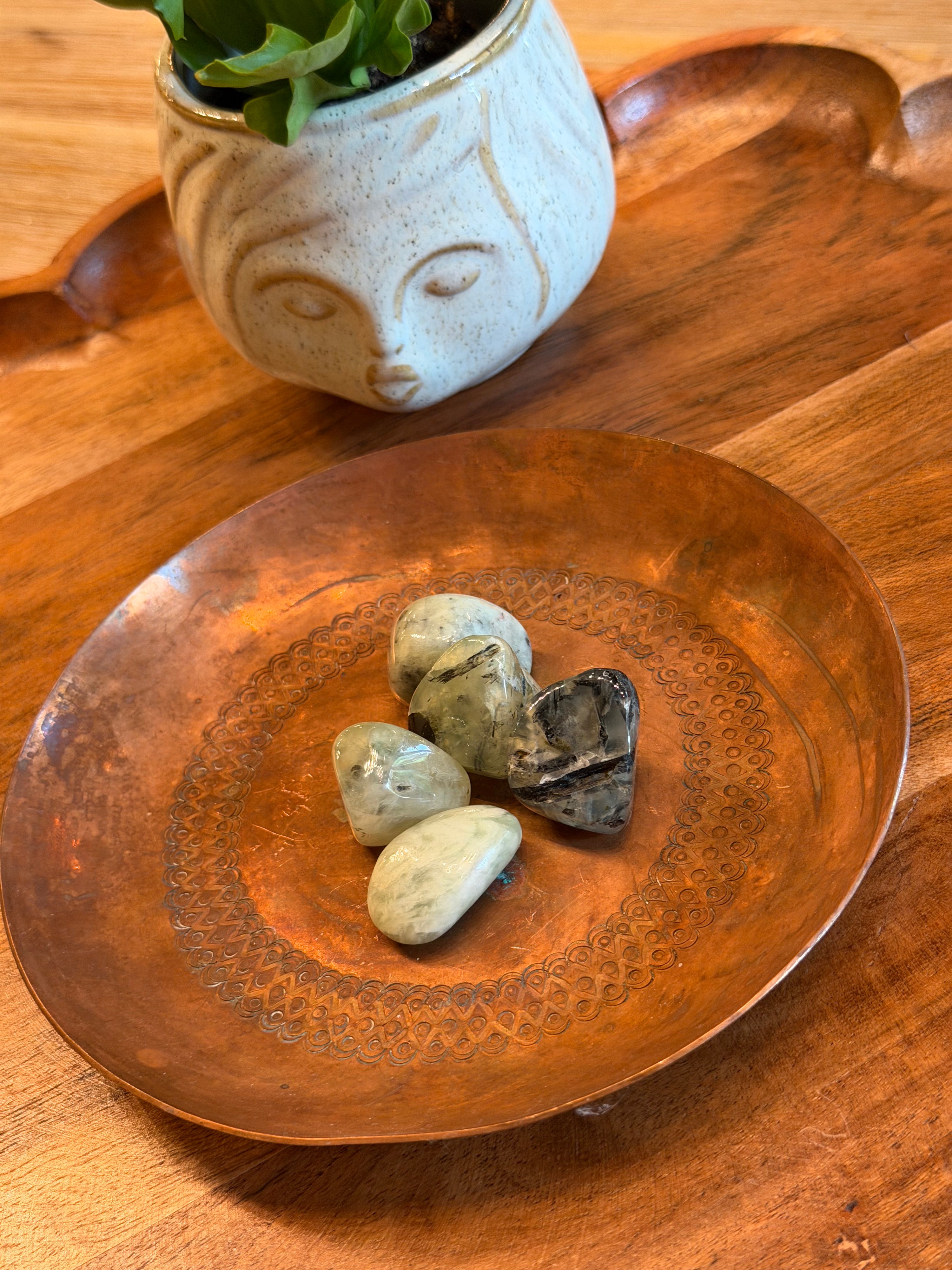 Four polished stones, including one heart-shaped stone, displayed on an engraved copper plate, with a small potted plant in the background on a wooden surface.