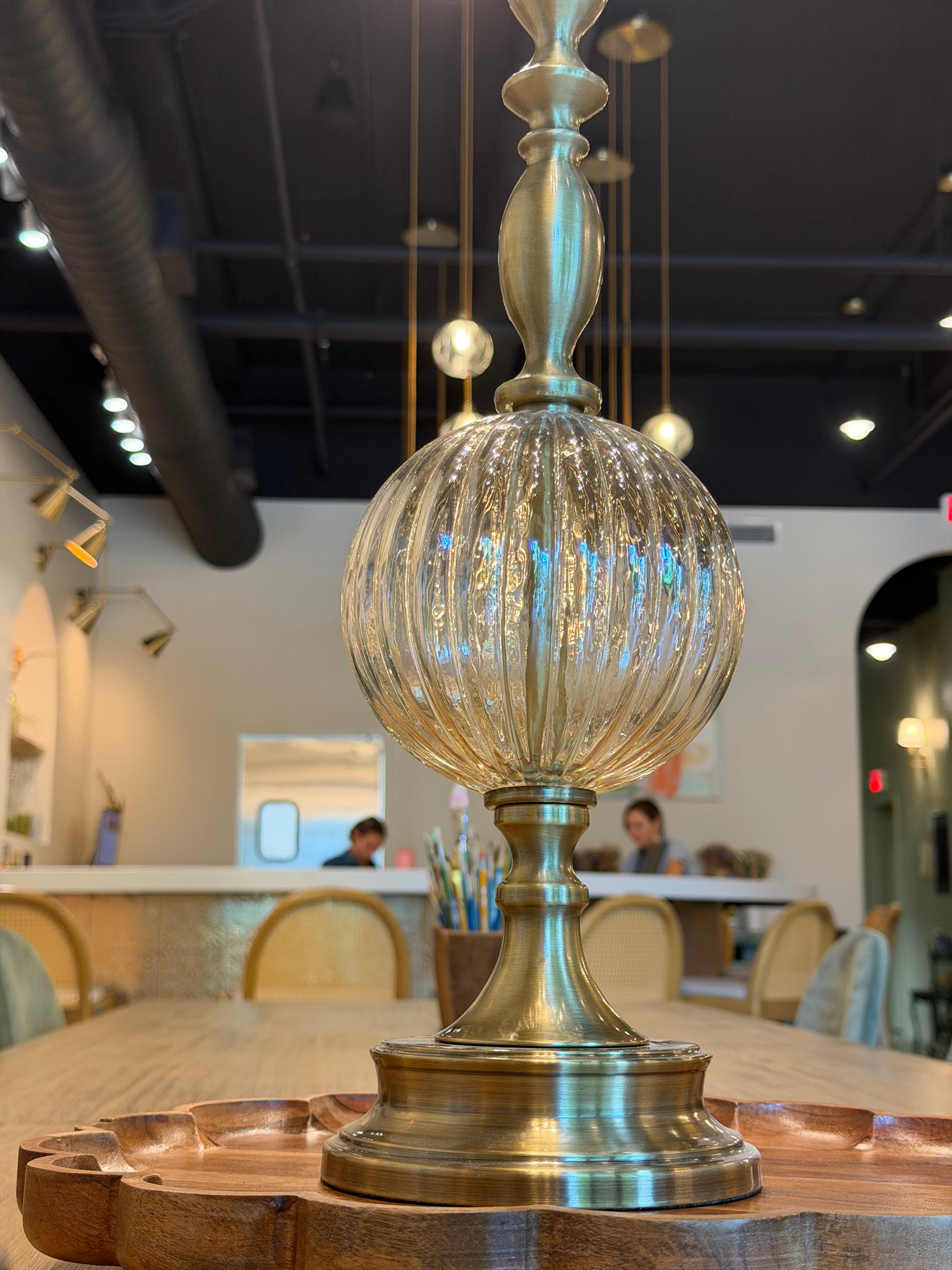 A decorative table lamp with a ribbed clear glass globe and a polished brass base, displayed on a wooden table in a modern cafe interior.