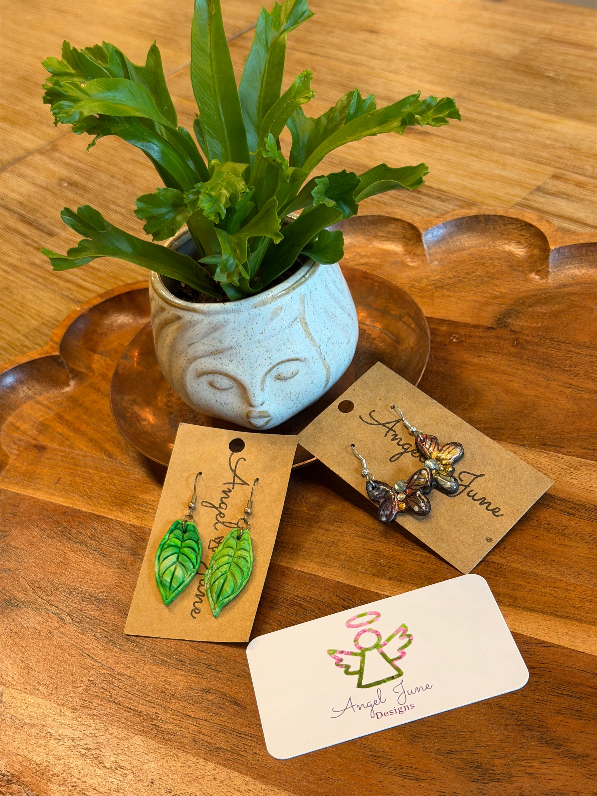 Three pairs of handmade earrings displayed on kraft and white cards on a wooden table, with a small potted green plant in the background. The cards show the brand name 'Angie Anne Earrings'.