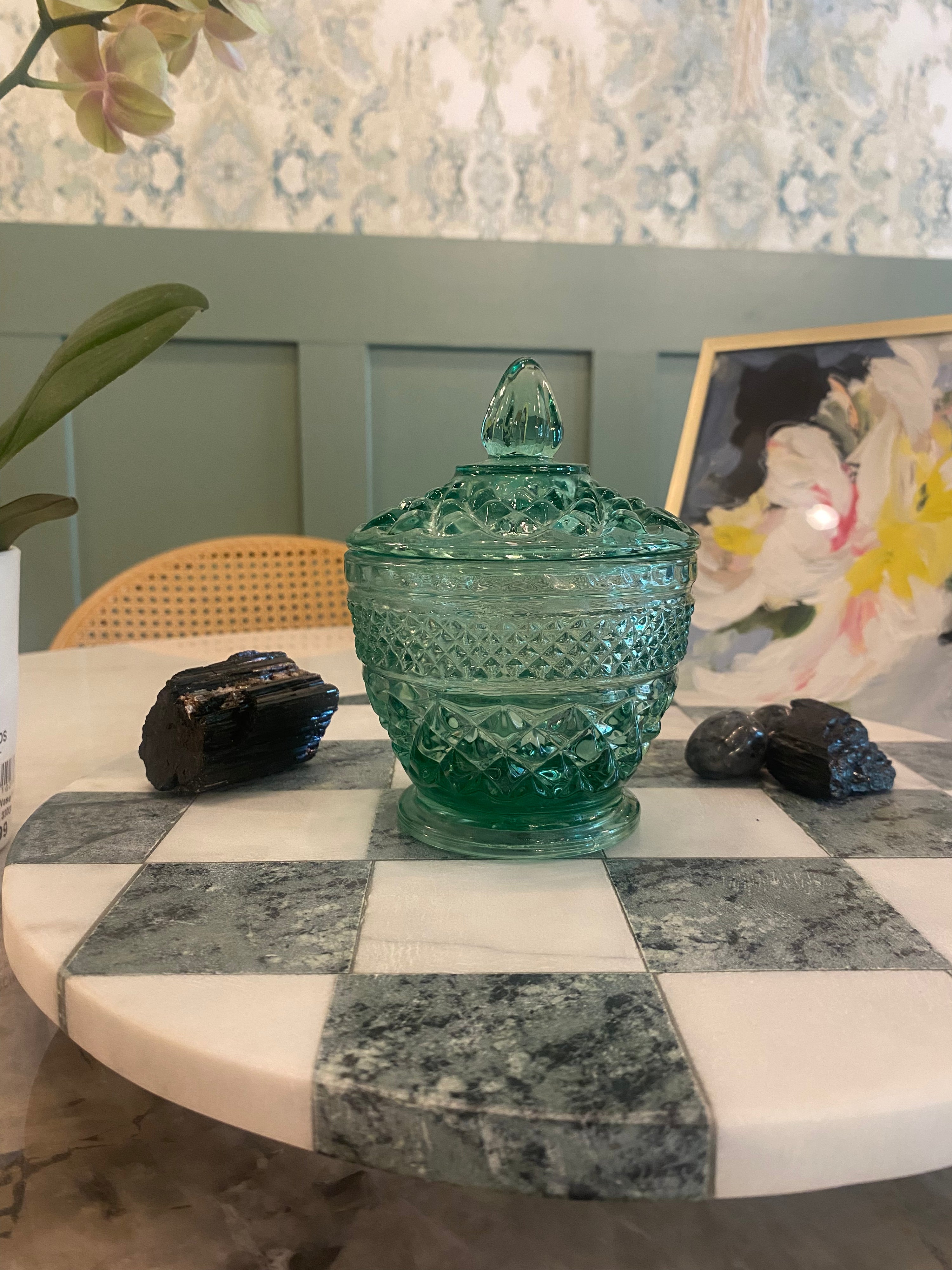 A vintage-style green glass lidded jar or candy dish with a diamond-cut pattern, displayed on a white and gray checkered marble table. The background shows a green paneled wall, a floral print, and some decorative rocks.