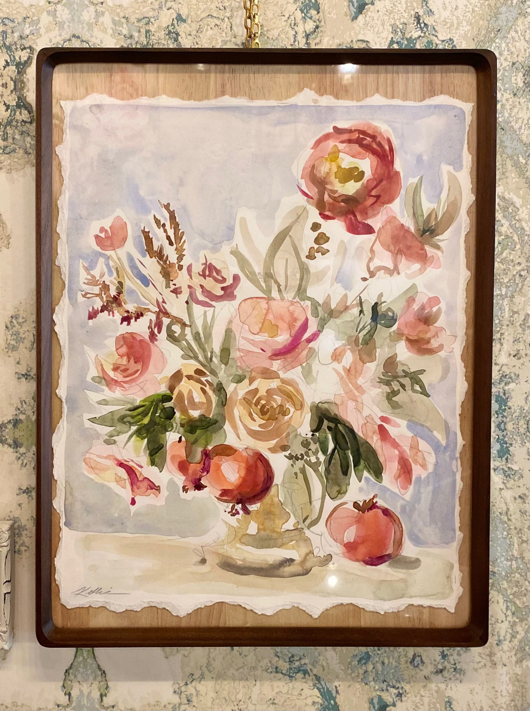 A framed watercolor painting of a floral bouquet with pomegranates, hanging on a textured wallpaper background. The painting features soft pink, red, and peach flowers with green leaves, set against a light blue-gray background, enclosed in a dark brown wooden frame.