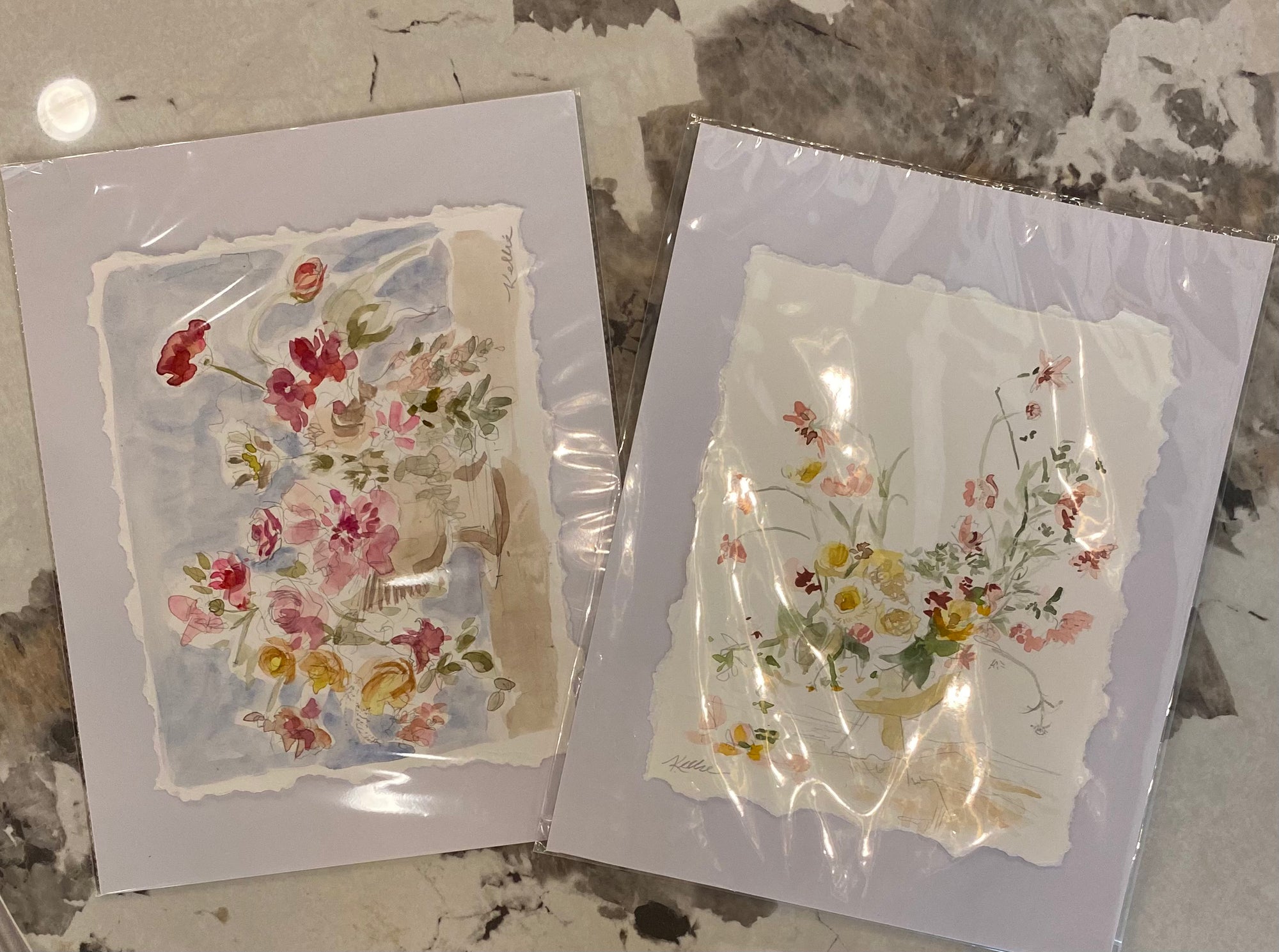 Two square floral art prints in clear plastic sleeves, displayed on a gray and white marble surface. The left print features a bouquet of red, pink, and yellow flowers on a light blue watercolor background, and the right print shows a bouquet of yellow, pink, and white flowers on a light beige background.