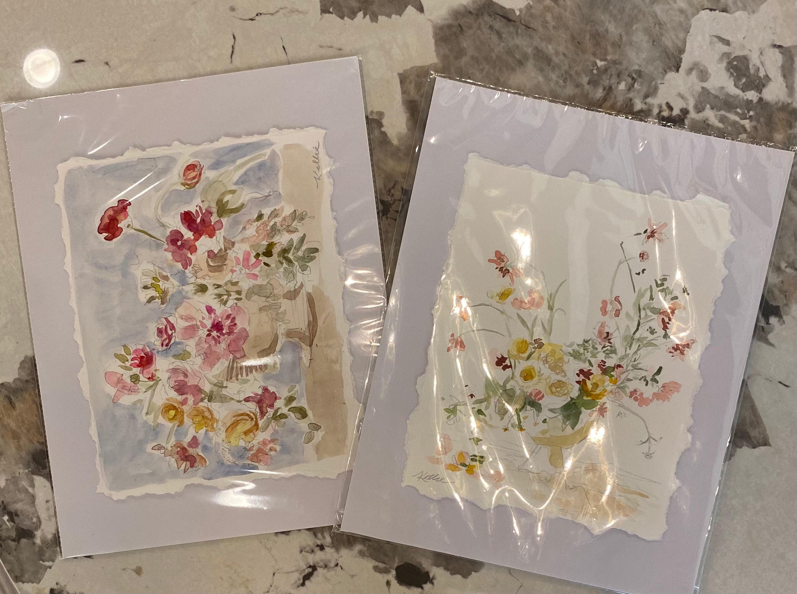 Two square floral art prints in clear plastic sleeves, displayed on a gray and white marble surface. The left print features a bouquet of red, pink, and yellow flowers on a light blue watercolor background, and the right print shows a bouquet of yellow, pink, and white flowers on a light beige background.