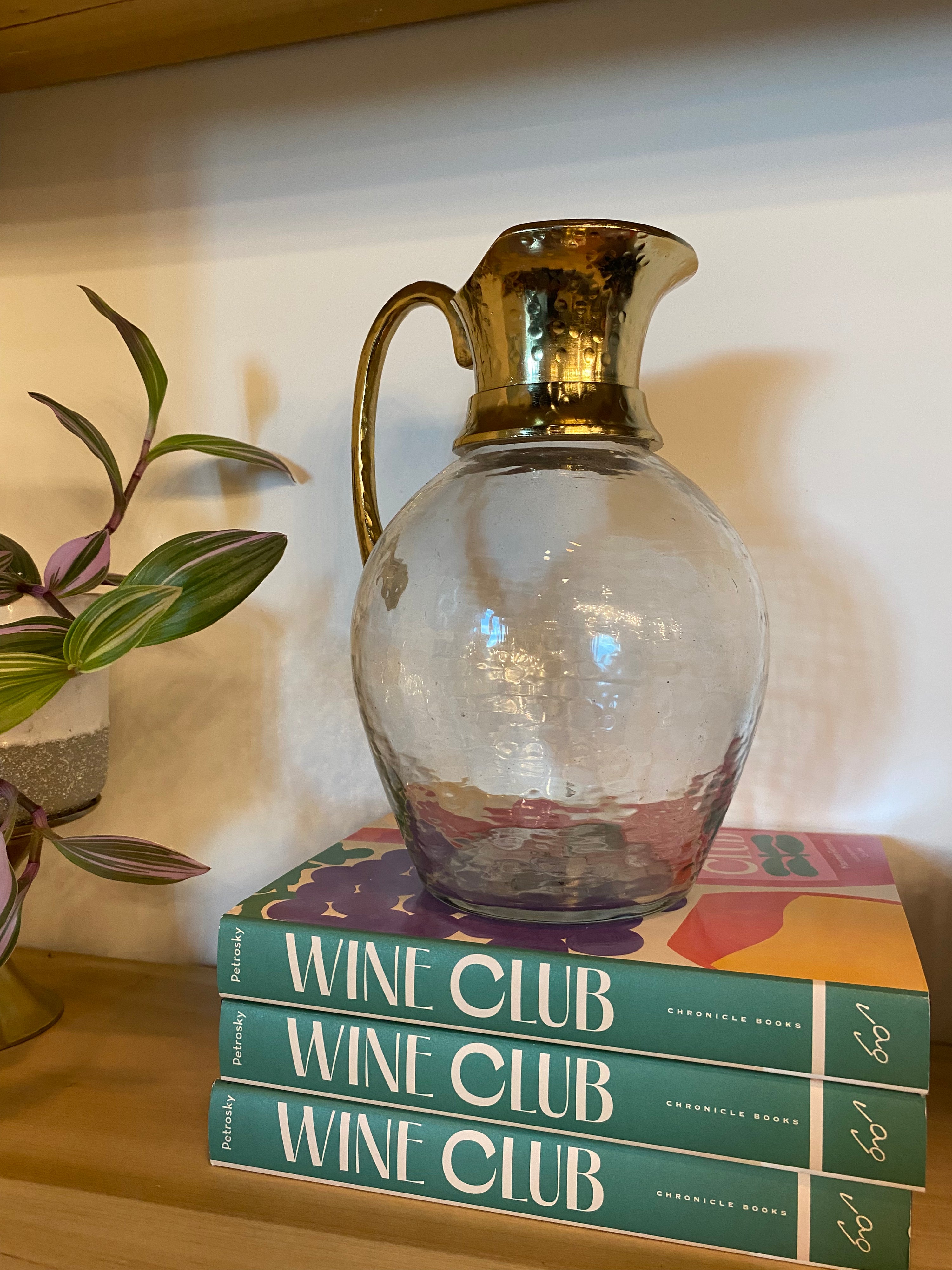 A clear glass pitcher with a hammered texture and a metallic gold-toned handle and spout, displayed on top of three books titled 'WINE CLUB' against a warm beige wall.
