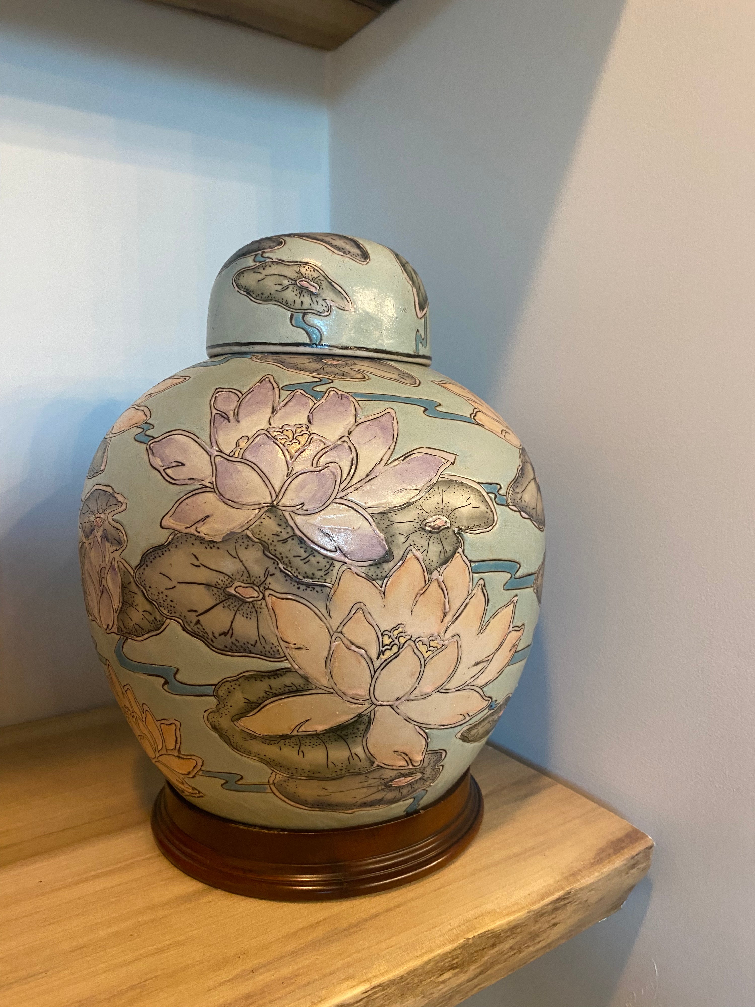 A ceramic tea jar with a lid, decorated with hand-painted lotus flowers and lily pads on a pale blue-green background, sitting on a wooden shelf.
