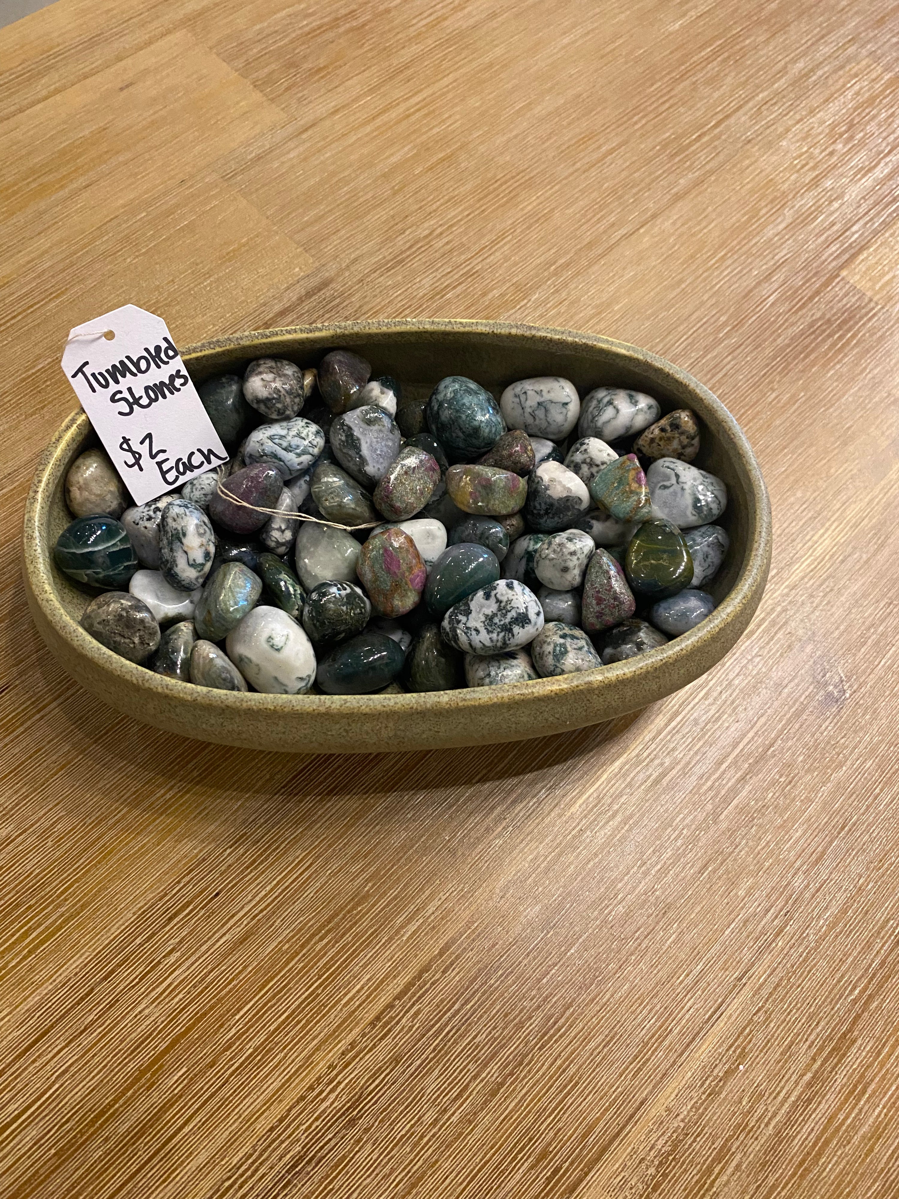 A bowl filled with polished tumbled stones, including a visible tag that reads 'Tumors Stones $2 Each,' placed on a wooden tabletop.